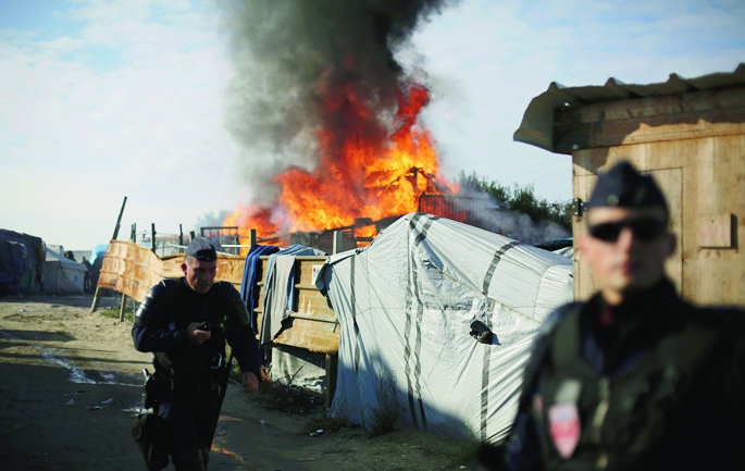 Riot police stand guard in front of a burning wooden house in the makeshift migrant camp known as "the jungle" near Calais, northern France, Tuesday, Oct. 25, 2016. Crews have started dismantling the migrant camp in France after the process to clear the camp began in earnest on Monday. (AP Photo/Thibault Camus) France Migrants