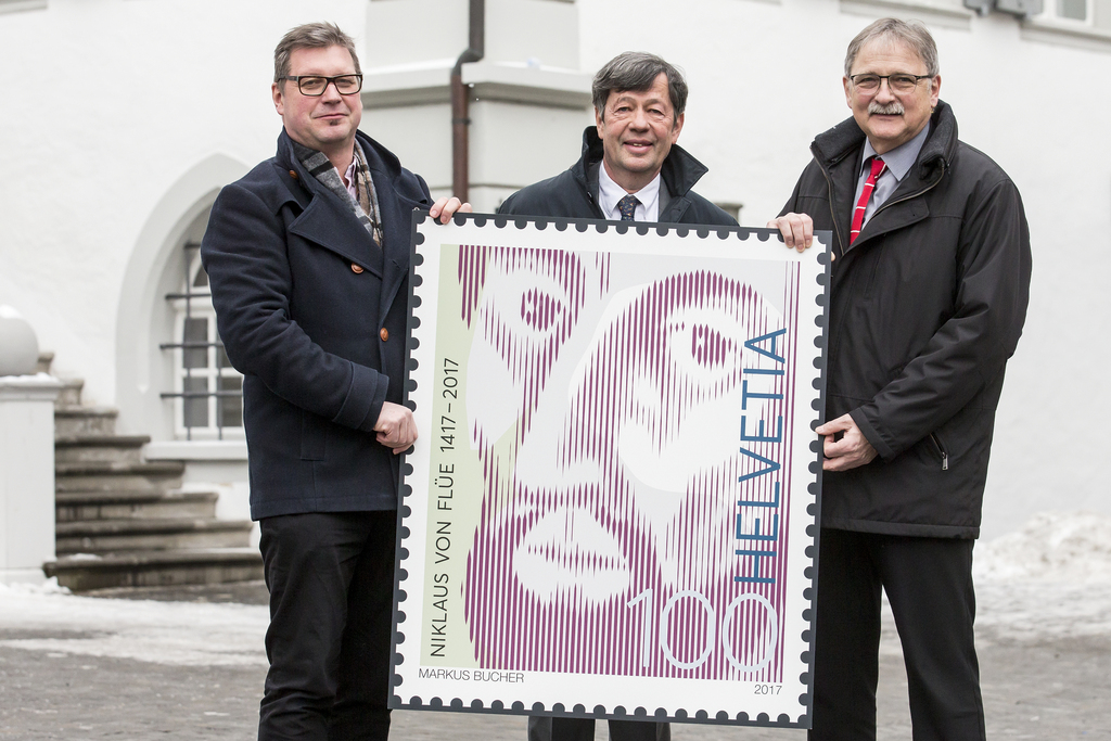 Le graphiste Markus Bucher, à gauche, Marco Durrer, de Poste Suisse, au centre, et Franz Enderli, président de l’association "600 ans Niklaus von Flüe" présentent le timbre de la commémoration.