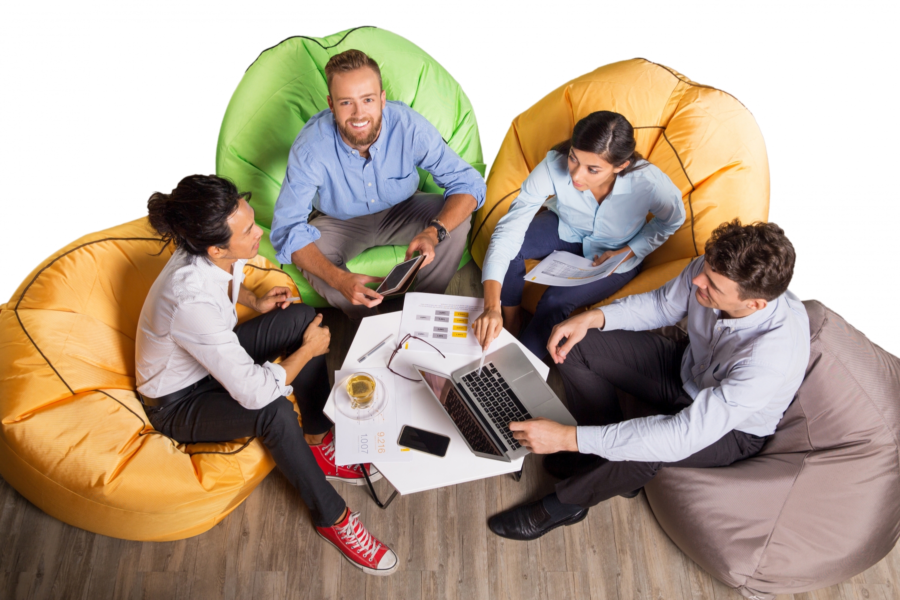 Four content young business people sitting on beanbag chairs around small café table, working and discussing issues. One man is looking at camera and smiling. High angle view.