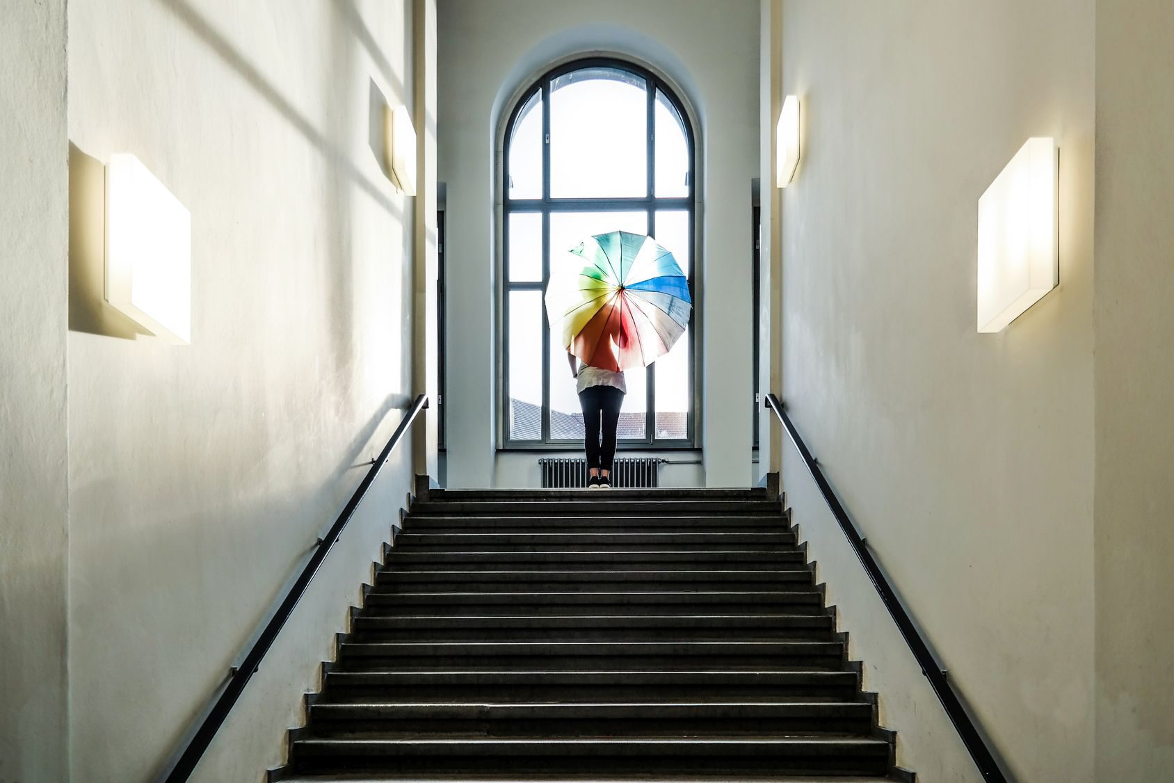 female with rainbow colored umbrella standing at top of stairs in front of window female with rainbow colored umbrella standing at top of stairs in front of window