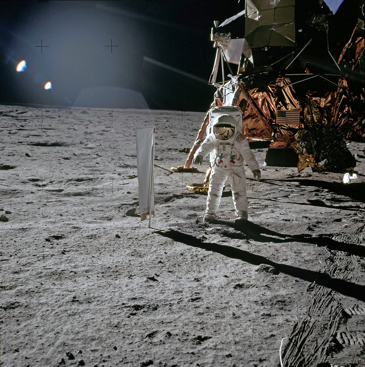 Astronaut Edwin E. Aldrin, Lunar Module (LM) pilot, stands beside the Solar-Wind Composition (SWC) Experiment,facing the camera. The LM is visible behind Adrin.  Linear trails (lines) in the right foreground were formed by the cable of the surface television camera.  The cable is visible on the lunar surface.  Image taken at Tranquility Base during the Apollo 11 Mission. Original film magazine was labeled S.  Film Type: Ektachrome EF SO168 color film on a 2.7-mil Estar polyester base taken with a 60mm lens. Sun angle is Medium. Tilt direction is Southeast (SE).
 Apollo 11 Mission image - Astronaut Edwin Aldrin stands beside t