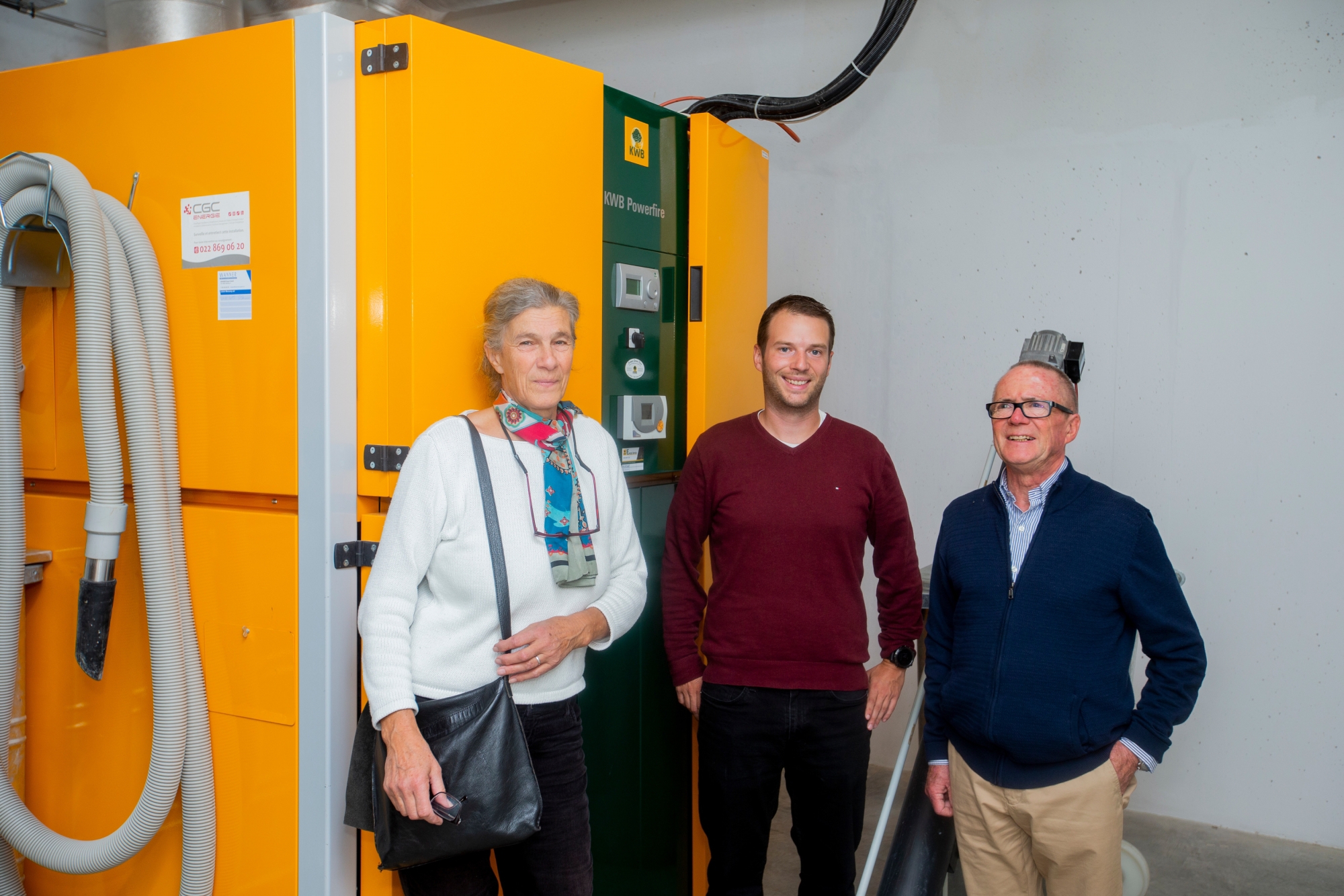  Marie-Béatrice Meriboute (maire de Céligny), Fabrice Baertschi (ingénieur) et Pierre Hoffmann (président de l'association ACP) posent devant la chaudière à bois.