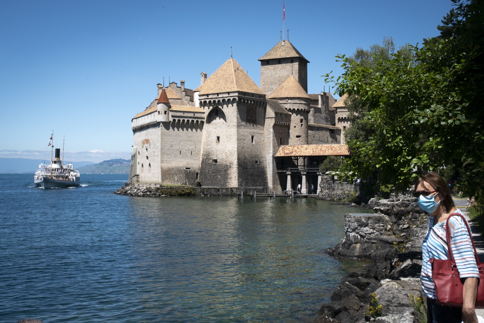 A tourist wearing protective face mask is pictured in front of the Chillon Castle (Chateau de Chillon) as the steamboat Vevey, CGN, sails on the Lake Geneva during the coronavirus disease (COVID-19) outbreak, in Veytaux near Montreux, Switzerland, Sunday, July 5, 2020. (KEYSTONE/Marcel Gillieron) ArcInfo